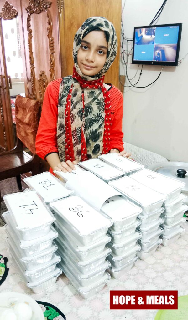 A young schoolgirl volunteer standing in front of meal boxes during Hope & Meals food distribution event on 26th September 2025.